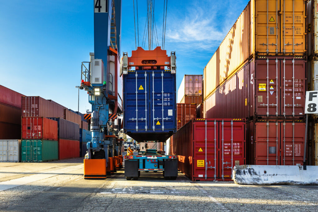 Ein blauer Container wird von einem Kranfahrzeug in einem Hafen auf die einen fahrbaren Untersatz gehoben-