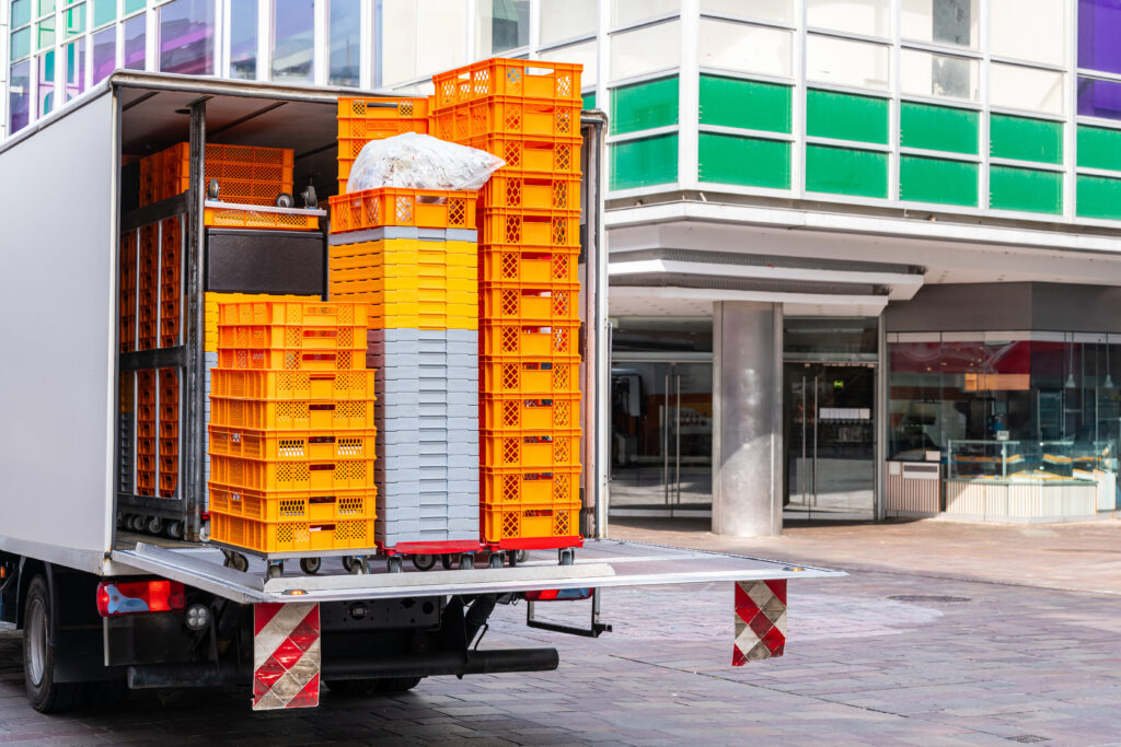 Lastwagen mit orangefarbenen und weißen Plastikboxen, die vor einem Geschäft ausgeladen werden.